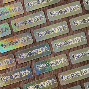 Zdjęcie przestawia ułożone równo brokatowe naklejki z napisem "smoczara". Naklejki mają zaokrąglone brzegi, a tło mieni się w kolorach tęczy.