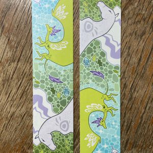 Zdjęcie przedstawia dwie zakładki do książki. Są one wydrukowane na perłowym, mieniącym się papierze. Na zakładkach widnieje jasnozielony smok z rozłożonymi skrzydłami oraz profil srebrzysto-liliowej klaczy. Tło stanowi błękitno-szmaragdowa mozaika.