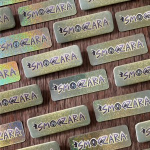 Zdjęcie przedstawia ułożone obok siebie brokatowe naklejki z napisem "smoczara" oraz prostokątne przypinki z takim samym napisem.